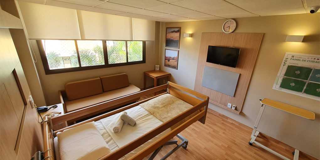 Suíte individual com cama hospitalar, sofá para acompanhante e janela ampla.